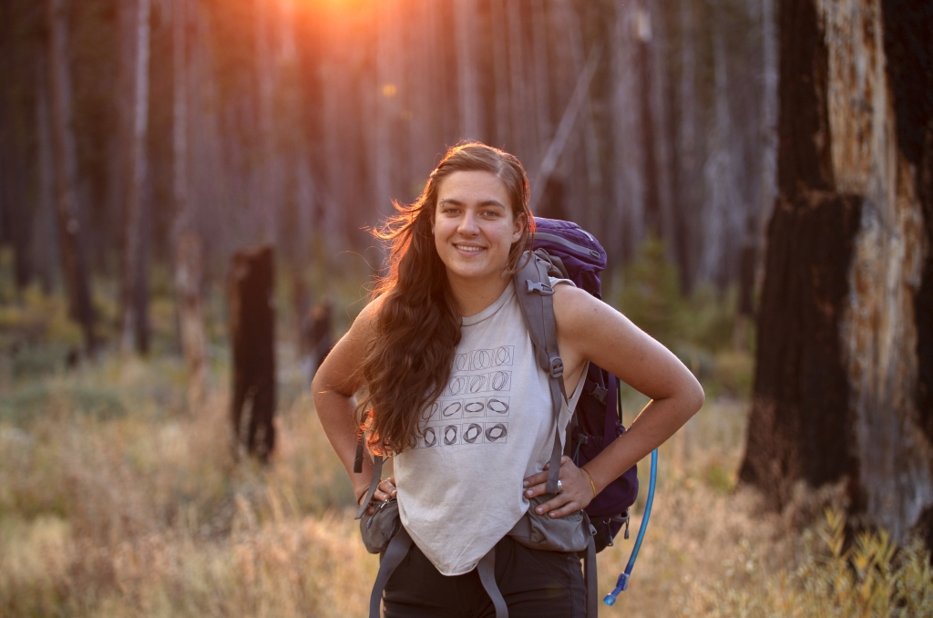 Woman stands among forest trees and sun sets.