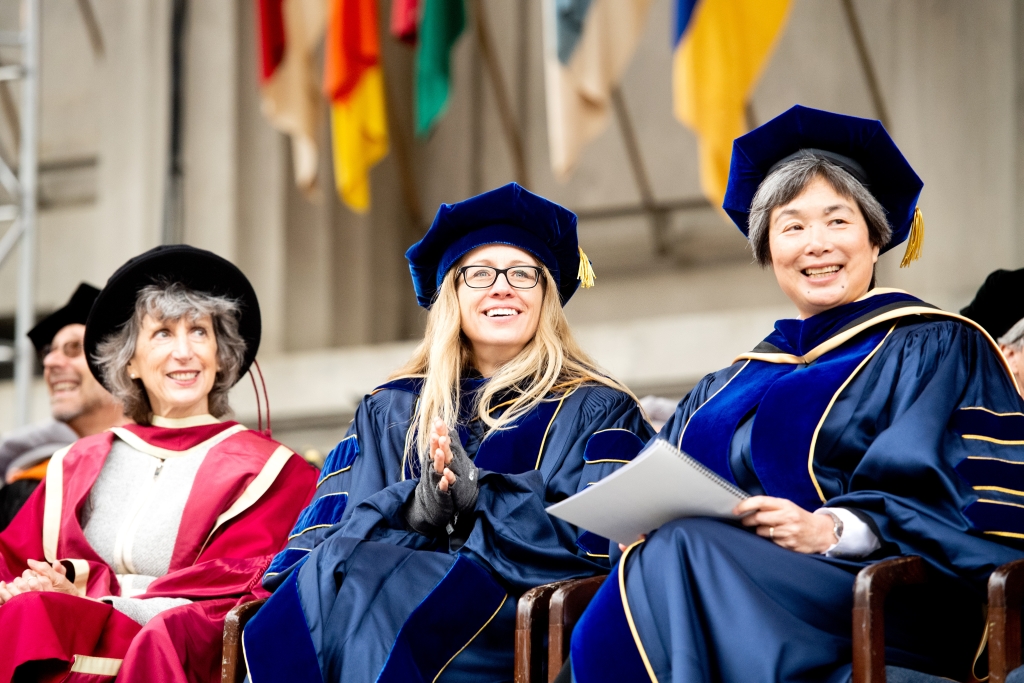 Three women in graduation regalia seats on stage.