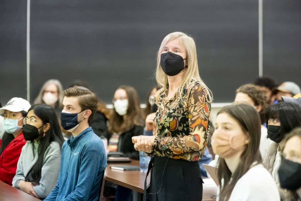 Seated students and standing professor look at screen located in foreground of photo.