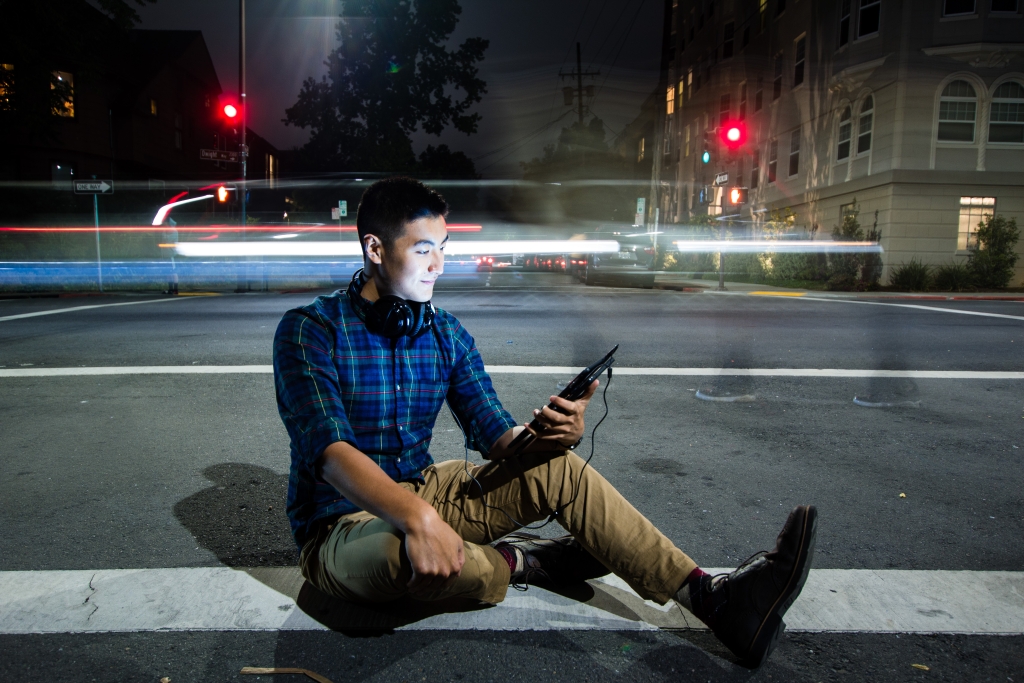 Man sitting on street with tablet and headphones, with street traffic in the background.