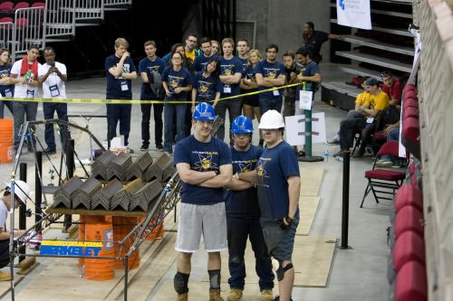 three male students pose beside steel bridge with hard hats on