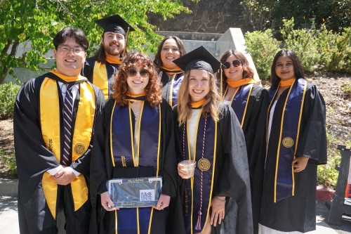 A group photo of graduating CEE students wearing graduation caps and gowns standing outside Davis Hall, smiling for a photo on O'Brien Breezeway. (Photo Credit: Pooja Nerkar/Berkeley CEE)
